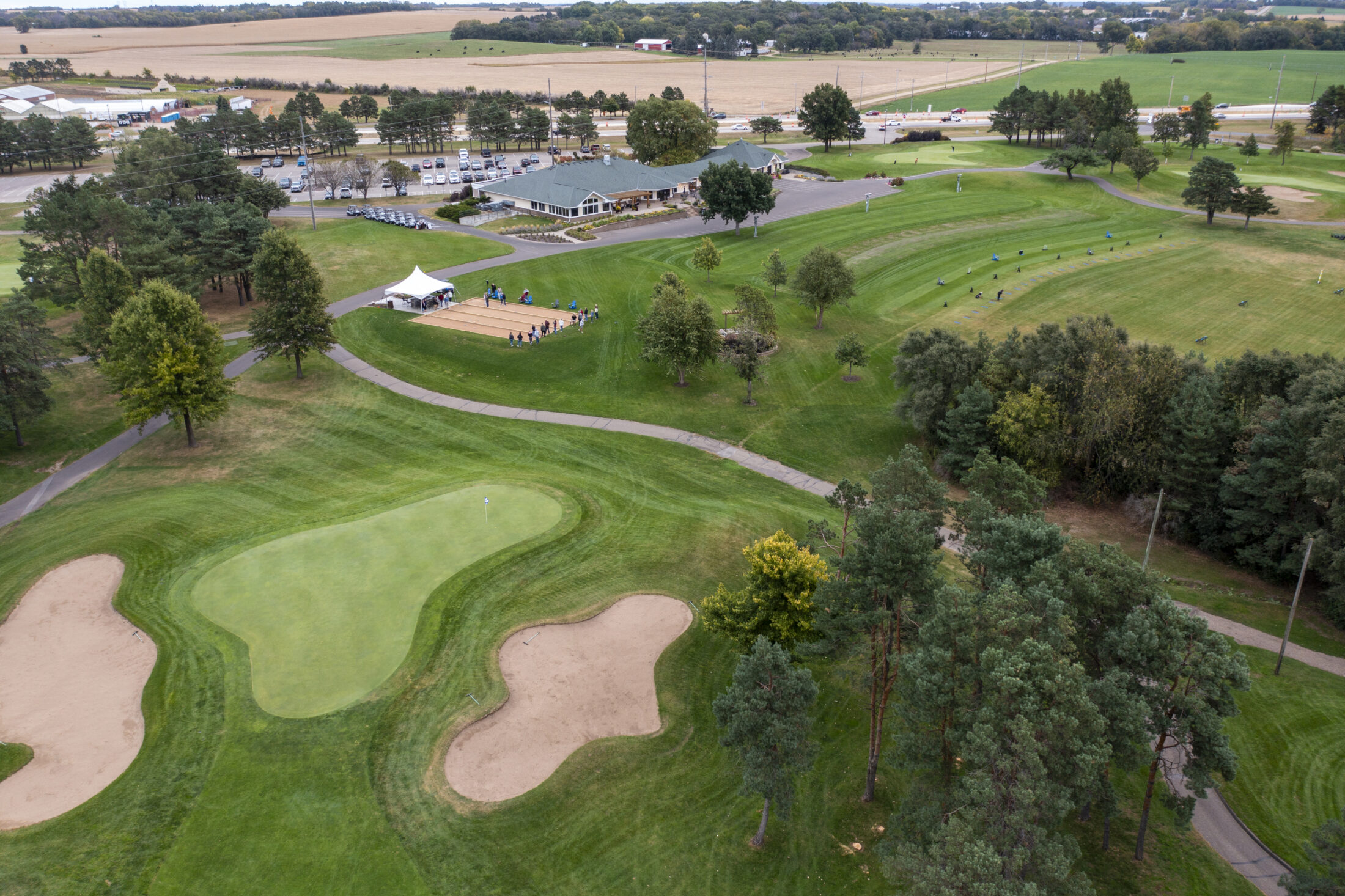 Aerial view of River Oaks Golf Course in Cottage Grove, Minnesota featuring greens, practice range, clubhouse, and outdoor event setup.