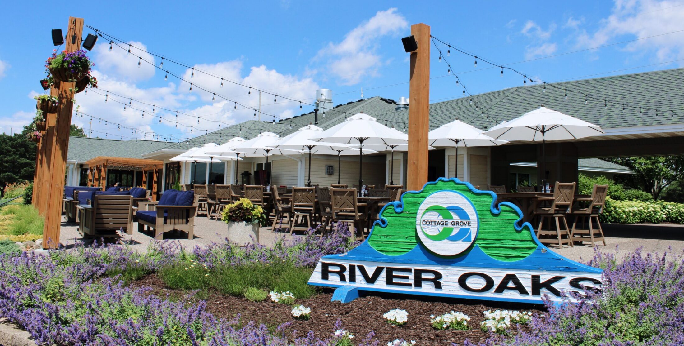 River Oaks Golf Course & Event Center entrance sign surrounded by seasonal flowers with the outdoor patio, umbrellas, string lights, and Eagles Bar & Grill in Cottage Grove, Minnesota.