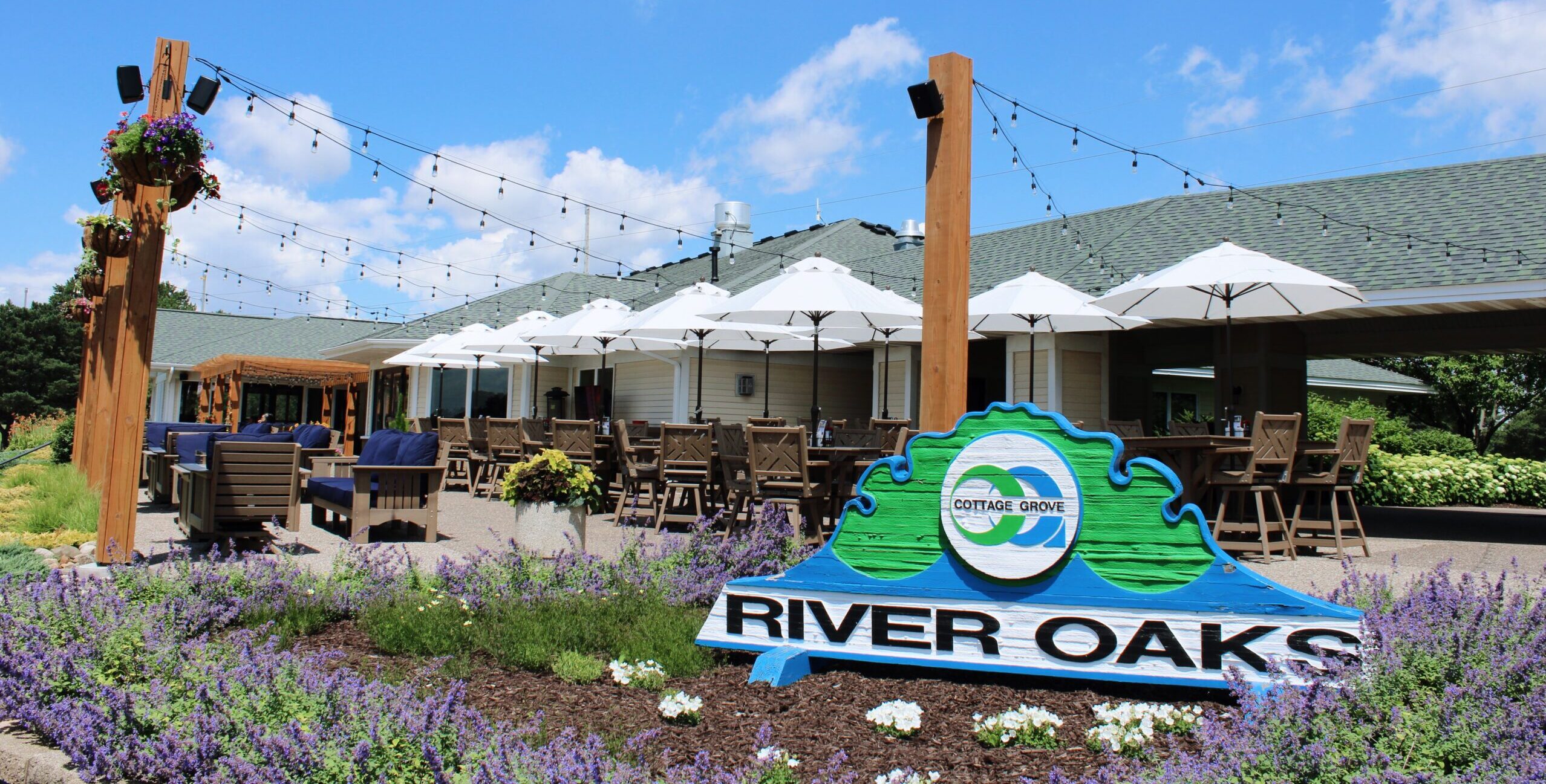 River Oaks Golf Course & Event Center entrance sign surrounded by seasonal flowers with the outdoor patio, umbrellas, string lights, and Eagles Bar & Grill in Cottage Grove, Minnesota.