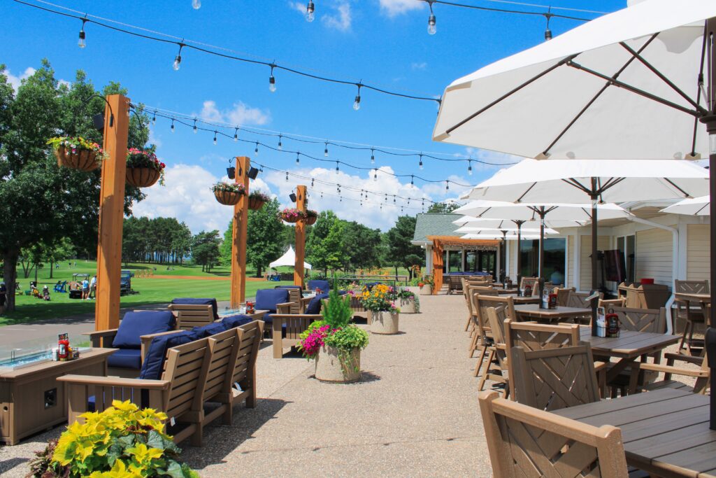 Outdoor patio at River Oaks Golf Course & Event Center featuring spacious dining tables with umbrellas, comfortable lounge seating, string lights, colorful landscaping, and scenic golf course views in Cottage Grove, Minnesota.