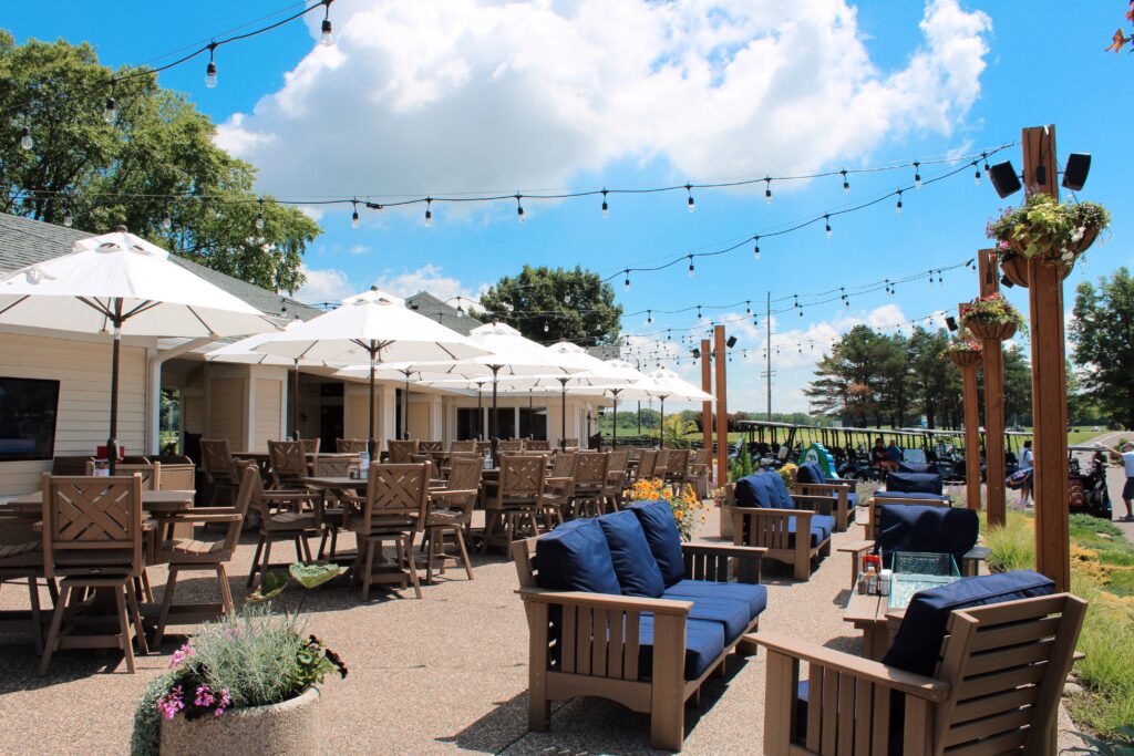 Outdoor patio at River Oaks Golf Course & Event Center with umbrella-covered dining tables, comfortable lounge seating, decorative string lights, and scenic golf course views in Cottage Grove, Minnesota.