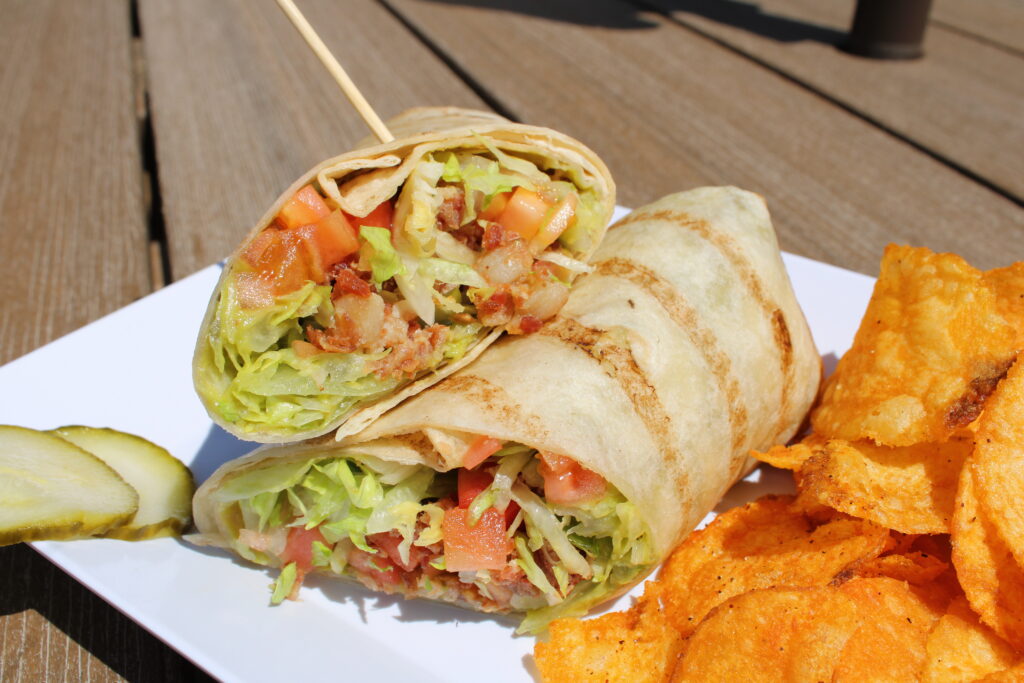 BLTA Wrap from Eagles Bar & Grill featuring bacon, lettuce, tomato, and avocado spread wrapped in a grilled tortilla and served with seasoned potato chips.