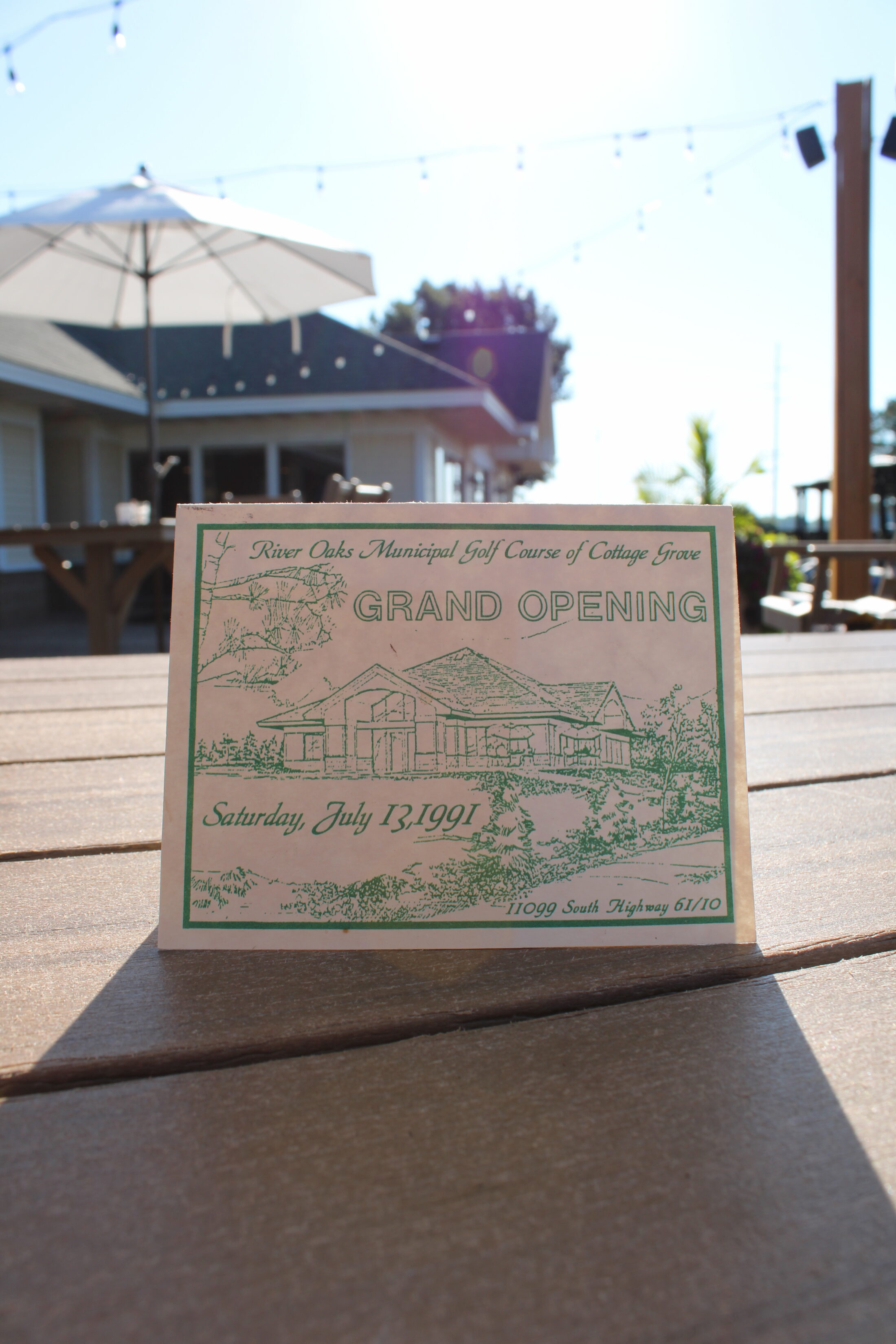 Historic River Oaks Municipal Golf Course grand opening invitation displayed on the River Oaks patio, commemorating the course's opening celebration on July 13, 1991, in Cottage Grove, Minnesota.